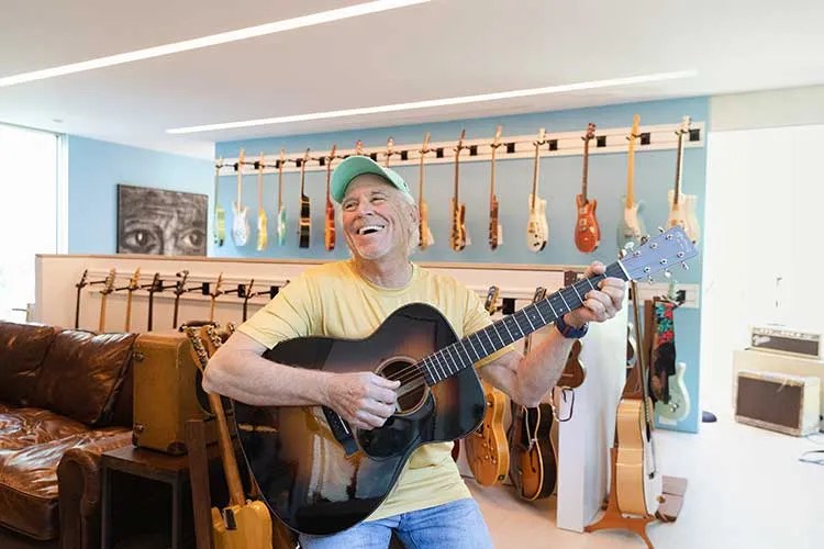 Jimmy Buffett smiling while playing a Martin acoustic guitar in his home
