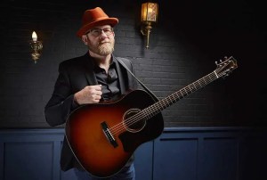 Bob Minner holding his guitar in front of a brick wall and lamp post
