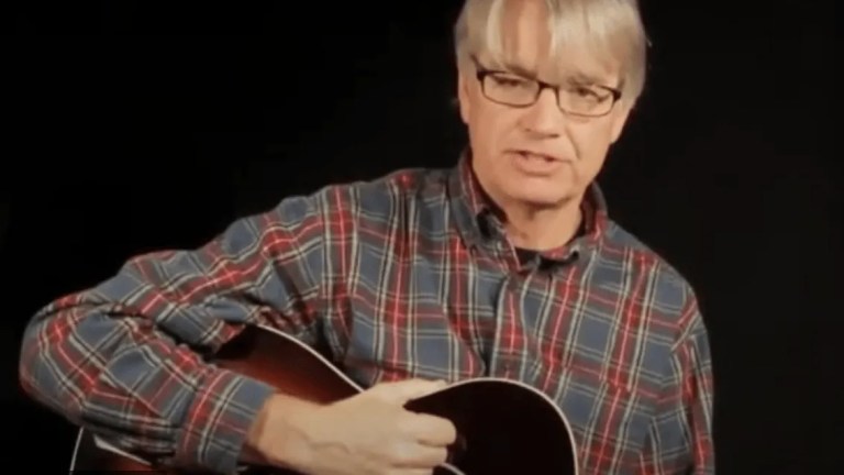 photograph of scott nygaard in a plaid shirt demonstrating a chord position on acoustic guitar